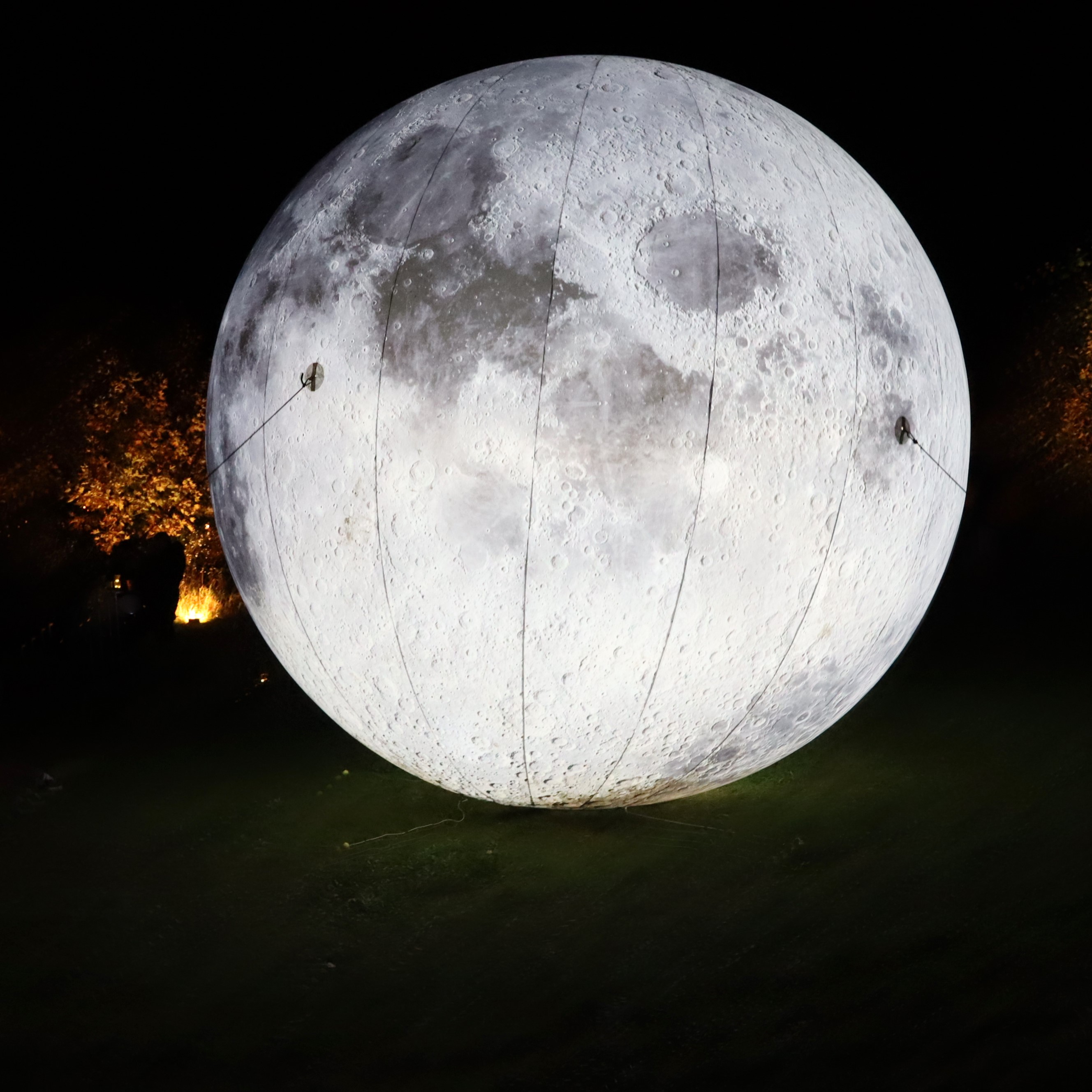 A large moon shines on the meadow in the city garden, with children in front of it and illuminated pylons in the background.